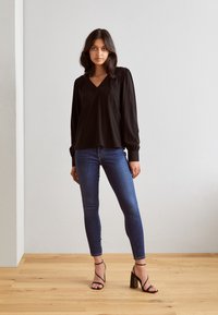 Blusa nera a maniche lunghe con scollatura a V, spalle arricciate, abbinata a jeans skinny blu e sandali con tacco a blocco neri e fasce.