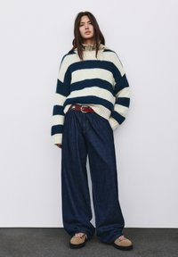 Donna che indossa un maglione a righe navy e bianche, jeans a gamba larga in denim scuro, stivali beige con lacci rosa e una cintura rossa, in piedi contro un muro bianco.