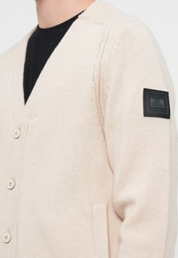 Light beige knitted cardigan with a V-neck, three buttons, ribbed cuffs, and a pocket. Features a branded black patch on the left sleeve.