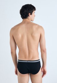 Zwarte zwemslip met een grijs gestreepte tailleband, met een strakke pasvorm en een gladde textuur, gezien vanaf de achterkant. Glad huid zichtbaar.