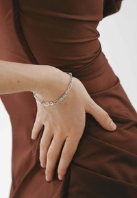 Pulsera de plata con formas ovaladas enlazadas y cinco piedras preciosas azules redondas, llevada en una mano que descansa sobre un fondo de tela marrón.