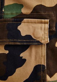 Close-up van een camouflage stof zak met klep, met groene, bruine, zwarte en tan onregelmatige vormen en zichtbare stiksels.