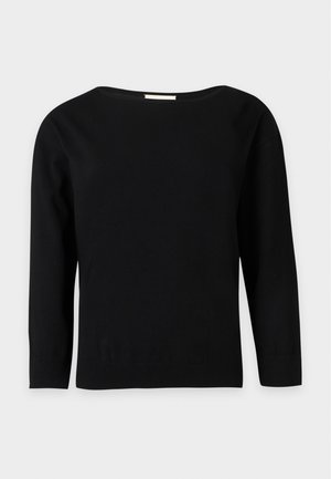 Femme aux cheveux courts et foncés portant un haut noir à manches longues, un pantalon blanc à jambes larges et des chaussures noires, debout devant un fond clair uni.