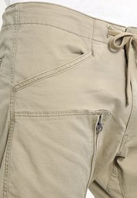 Pantaloni beige con molte tasche, tra cui una tasca con zip e una vita con cordino, indossati con una camicia bianca.