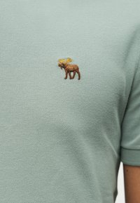 Φωτεινό πράσινο υφασμάτινο με υφή, με μικρό κεντημένο καφέ ελάφι με κίτρινα κέρατα στην περιοχή του στήθους ενός ενδύματος με κοντά μανίκια.