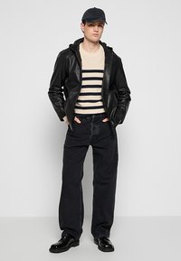Giacca di pelle nera sopra un maglione a righe crema, pantaloni in denim scuro e scarpe nere. Il modello indossa un berretto scuro. Design semplice e casual.