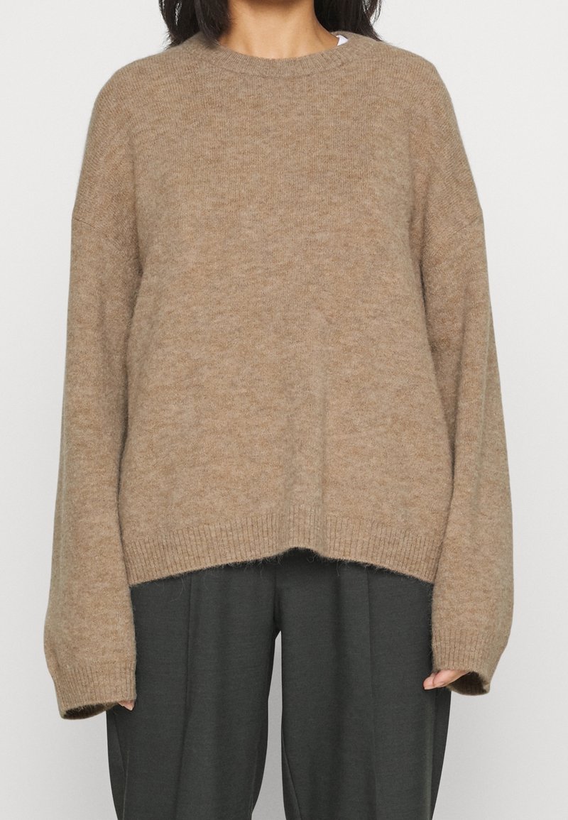 Femme portant un pull en maille beige ample avec des épaules tombantes et des manches longues, associé à un pantalon large gris foncé.