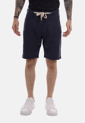 Uomo con pantaloncini blu navy con cintura in corda bianca, camicia nera, sneakers bianche e tatuaggi su entrambi gli avambracci, in piedi con le braccia rilassate.