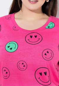 Camiseta rosa con varios diseños de caras sonrientes en negro y verde, con un escote redondeado y una textura de tela suave y lisa.