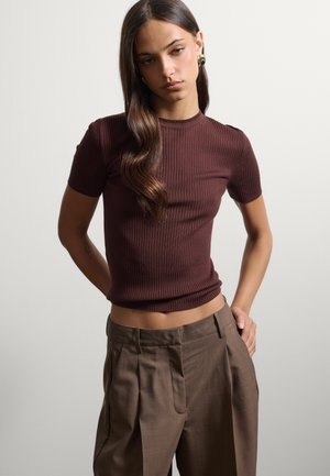 Femme aux longs cheveux bruns portant un haut marron côtelé à manches courtes et ajusté, ainsi qu'un pantalon marron ample taille haute, sur un fond uni.