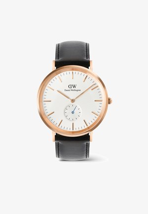 Daniel Wellington óra fehér textúrált számlappal, rózsaarany tokban, fekete bőrszíjjal és egy kis kék szubszámlappal az alján.