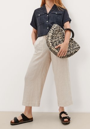 Chemise cropped marine à manches retroussées, pantalon en lin beige à jambes larges et sac noir tissé avec poignée. Sandales noires avec boucles et semelles tissées.