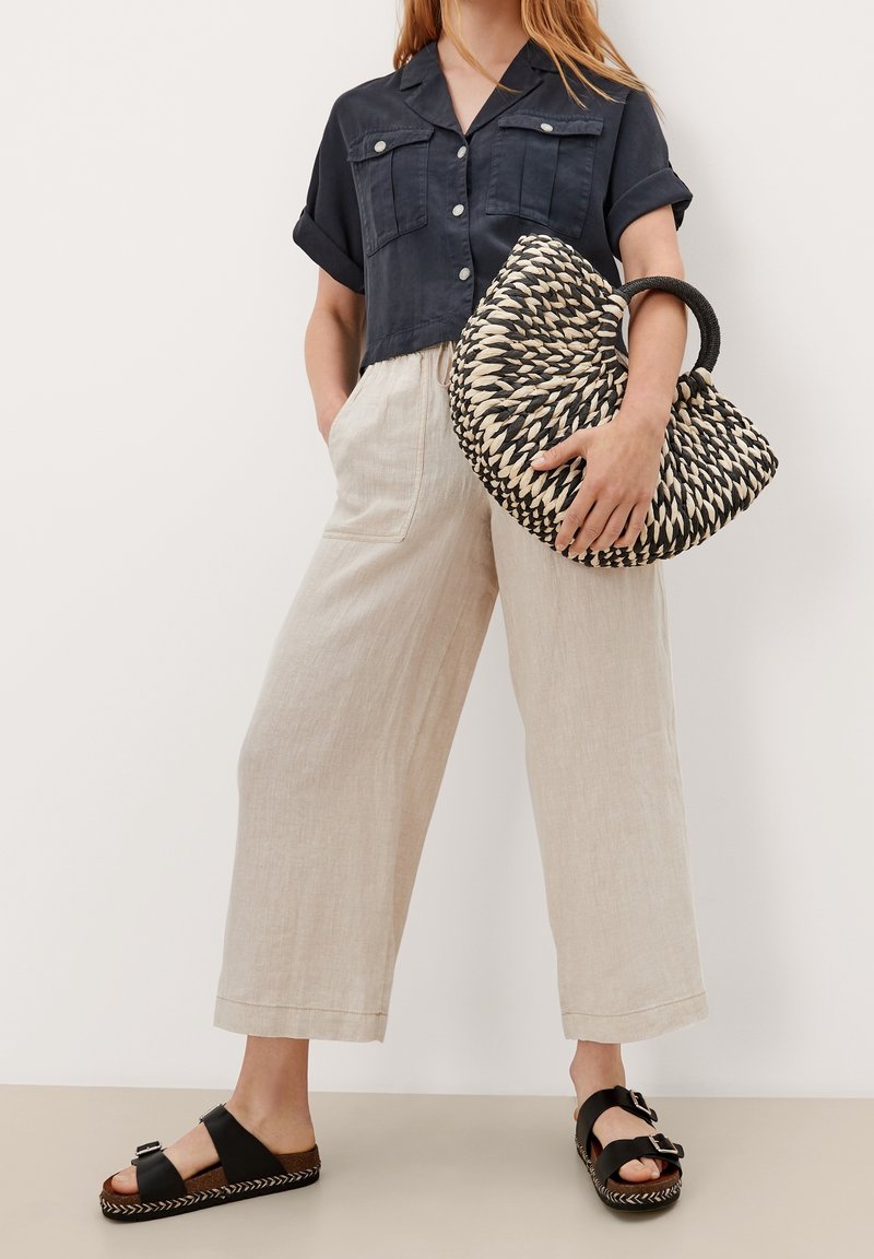 Camicia blu navy accorciata con maniche arrotolate, pantaloni larghi in lino beige, e borsa nera intrecciata con manico. Sandali neri con fibbie e suole intrecciate.