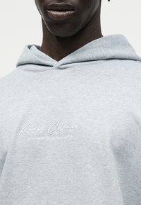 Άτομο με σκουρόχρωμο δέρμα που φοράει ανοιχτογκρι φούτερ με κουκούλα και λευκό κεντητό κείμενο "Carlo Colucci A Legacy Since 1978" στο στήθος.