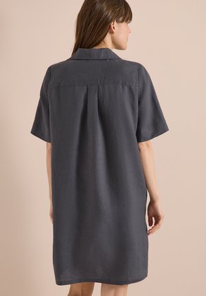 Femme aux cheveux bruns portant une robe ample à manches courtes gris foncé, debout de dos sur un fond beige.