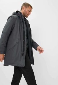 Uomo che cammina indossando un cappotto invernale grigio scuro con cappuccio sopra pantaloni neri, su uno sfondo chiaro e uniforme.