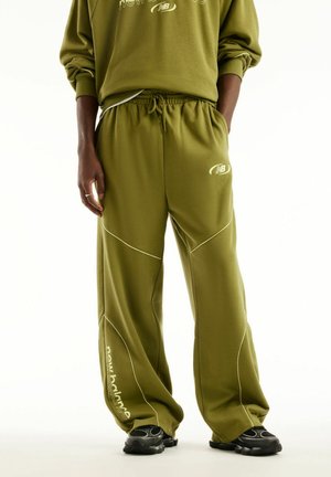 Persona che indossa pantaloni della tuta verde oliva New Balance e felpa abbinata, in piedi con le mani nelle tasche e scarpe da ginnastica nere.