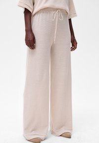 Pantalons en tricot beige à taille haute et jambes larges avec cordon de serrage, portés avec un haut assorti et des chaussures beiges à bout pointu sur un modèle à la peau noire.