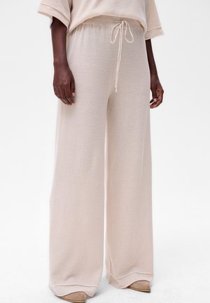 Pantalons en tricot beige à taille haute et jambes larges avec cordon de serrage, portés avec un haut assorti et des chaussures beiges à bout pointu sur un modèle à la peau noire.