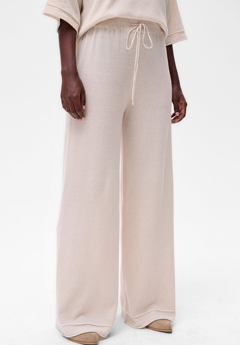 Beige, gebreide broek met wijde pijpen en hoge taille, met een trekkoord, gedragen met een bijpassend top en beige, puntige schoenen op een donkerhuidig model.