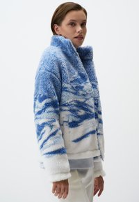 Giacca fuzzy, blu e bianca con motivo a onde, colletto alto, chiusura con zip e texture morbida, caratterizzata da design ondulati che richiamano l'acqua.