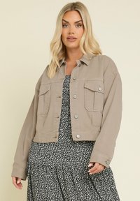 Beige denimjacka med knapppning, två framfickor och långa ärmar, lager på lager över en svart blommig klänning.