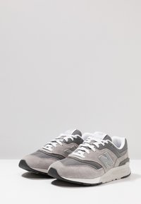 Tenisi New Balance gri, realizati dintr-un amestec de piele intoarsa si material mesh. Sireturi albe si branding pe limba si pe lateral, cu accente argintii.