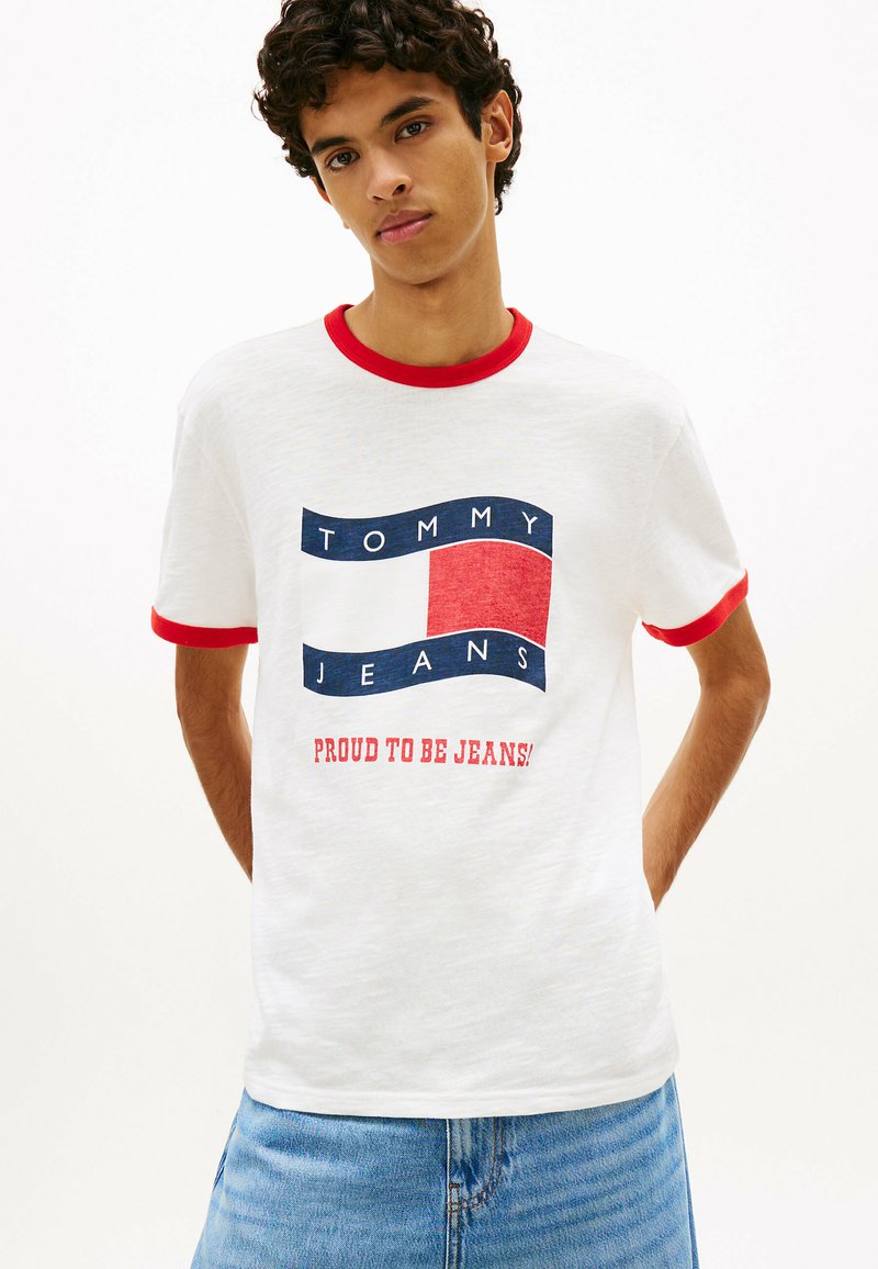 Jeune homme aux cheveux bouclés portant un t-shirt blanc avec bordure rouge et le logo "Tommy Jeans Proud to Be Jeans!", associé à un jean en denim bleu clair.