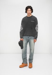 Joven se encuentra de pie contra un fondo blanco, vistiendo un sudadera negra desgastada con diseños en las mangas en blanco, jeans claros desgastados y botas beige, sosteniendo una gorra negra.