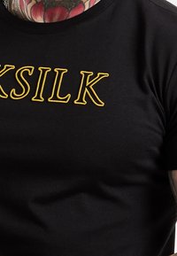 Mustapaita, jossa on suuri keltainen brodeerattu teksti "SILK". Kangas on sileä ja siinä on klassinen pyöreä kaula-aukko.