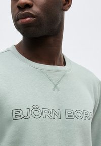Κοντινό πλάνο ενός ατόμου που φοράει ένα ανοιχτό πράσινο φούτερ με την επιγραφή "BJÖRN BORG" τυπωμένη με μαύρα γράμματα στο στήθος.