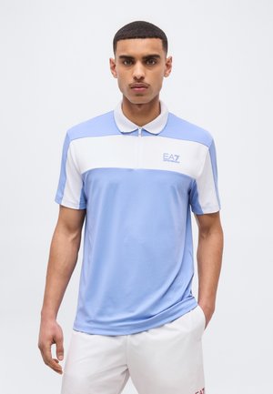 Νεαρός άνδρας στέκεται με τα χέρια στις τσέπες, φορώντας μπλε ανοιχτό και λευκό πόλο με το λογότυπο EA7, και λευκό παντελόνι, σε απλό φόντο.