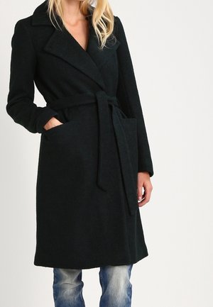 Femme portant un long manteau noir ceinturé avec col à revers et poches, associé à un jean bleu, sur un fond blanc uni.