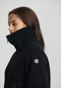 Zwart fleecejack met hoge kraag, gestructureerd materiaal, zijzakken met rits en een ronde logo patch op de linker mouw.