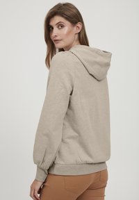 Kapa s puloverjem v svetlo bež barvi, s sproščenim krojem, rebrastimi manšetami in mehkobo teksture. Očitne podrobnosti šivanja in elastični pas.