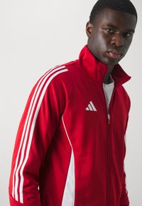 Rød atletisk jakke med høj krave, hvide striber langs ærmerne og lynlås lukning. Adidas-logo i hvidt på venstre bryst. Glat stoftekstur.