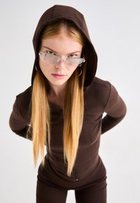 Sudadera con cremallera marrón hecha de tela suave y con una capucha detallada. Gafas claras e innovadoras con acentos plateados y aros estampados.