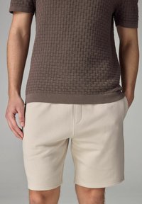 Chemise marron à manches courtes avec une texture, ornée d'un motif en carreaux, associée à un short beige clair au design simple et aux poches avant.