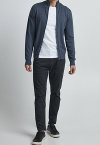 Mężczyzna w granatowym rozpinanym swetrze na białej koszulce, czarnych spodniach i czarnych sportowych butach na tle jednolitego jasnego tła.