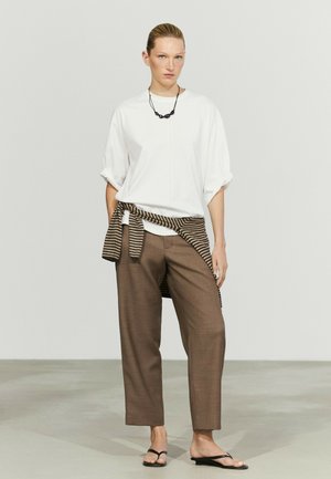 Donna con camicia bianca larga, pantaloni marroni corti, collana nera, maglione a righe legato in vita e sandali infradito neri in piedi.