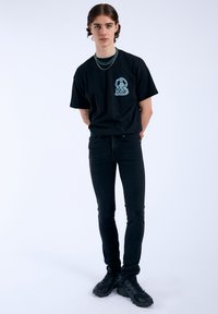 Camiseta negra de manga corta con un gráfico azul intrincado en el pecho, combinada con jeans ajustados negros y zapatos negros gruesos.
