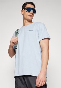 Světle modré bavlněné tričko s krátkým rukávem a kulatým výst�řihem. Na hrudi má malé logo Patagonia. Model drží popruh batohu.