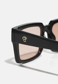 Ochelari de soare negri cu un design unghiular, lentile maro tinted și un mic logo galben pe braț. Finisaj din plastic neted și lucios.