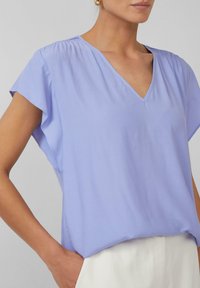 Licht lavendelblouse met v-hals en korte, fladderende mouwen. Gemaakt van zacht, soepel stof met subtiele plooien op de schouders.