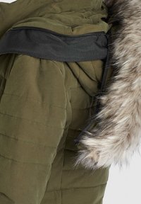 Olivgrön puffjacka med dragkedjedetalj vid övre axel och fuskpäls längs huven. Kviltat mönster och platt svart elastiskt band.