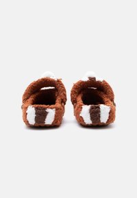 Καφέ και λευκές χνουδωτές παντόφλες με κλειστή φτέρνα, σχεδιασμένες για εσωτερική χρήση, εμφανίζονται από πίσω σε απλό λευκό φόντο.