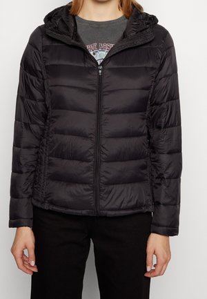 Personne portant une veste matelassée noire zippée avec capuche, un t-shirt gris à motif et un pantalon noir, debout devant un fond uni.