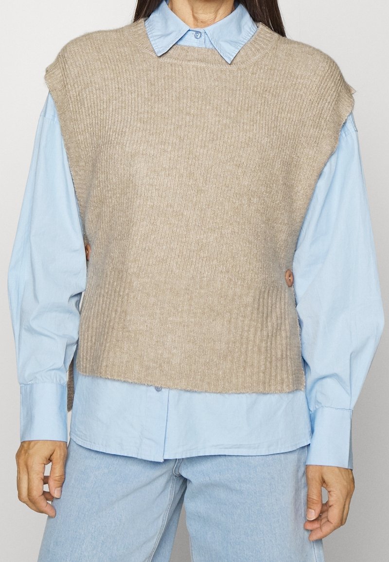 Femme portant un gilet en maille sans manches beige sur une chemise boutonnée bleu clair et un jean bleu clair, debout devant un fond uni.