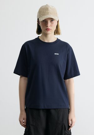 Νεαρό άτομο που φοράει μπεζ καπέλο BOSS, μπλε ναυτικό T-shirt BOSS και μαύρο παντελόνι cargo, στέκεται μπροστά σε απλό φωτεινό φόντο.