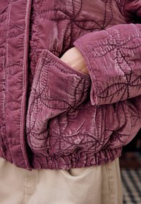 Steppjacke aus mauvefarbenem Samt mit strukturiertem Muster, ausgestattet mit einem Bündchenärmel und einer seitlichen Tasche. Der Stoff hat einen weichen Glanz und eine geraffte Taille.